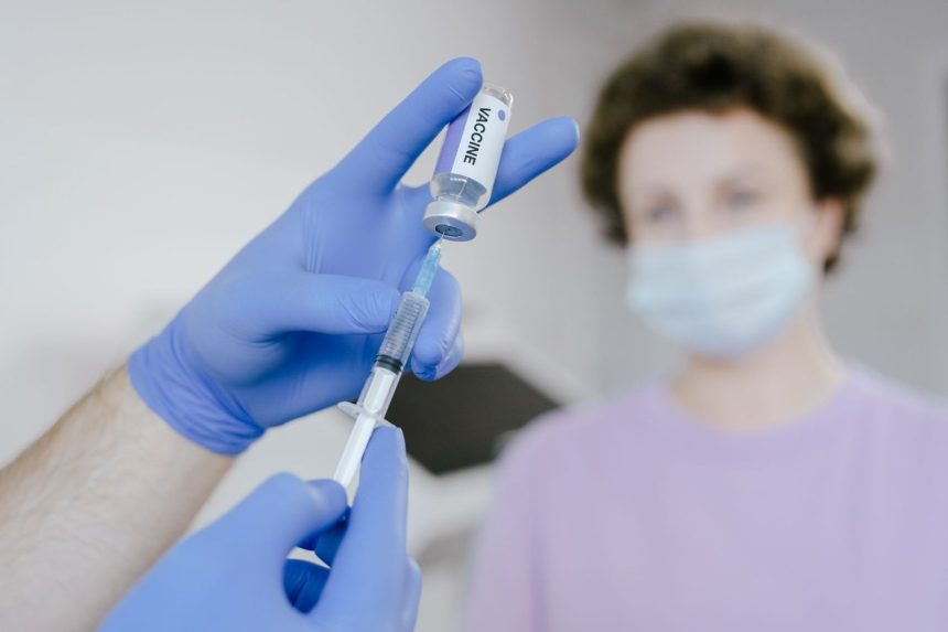 Profissional de saúde, usando luvas azuis, prepara uma seringa com vacina enquanto um paciente aparece ao fundo.