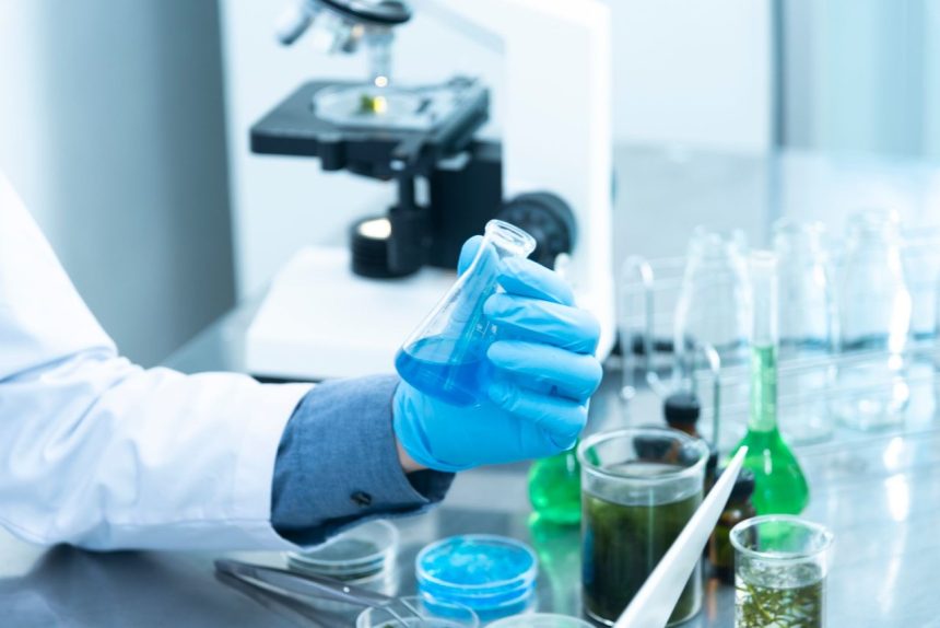Cientista usando luvas analisa uma amostra azul em laboratório, com microscópio e vidrarias ao fundo.