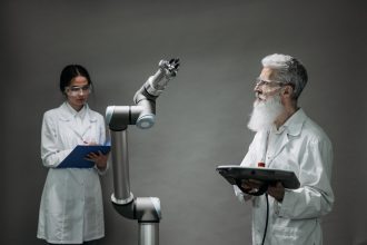 Dois pesquisadores de jaleco analisam um braço robótico em um laboratório.