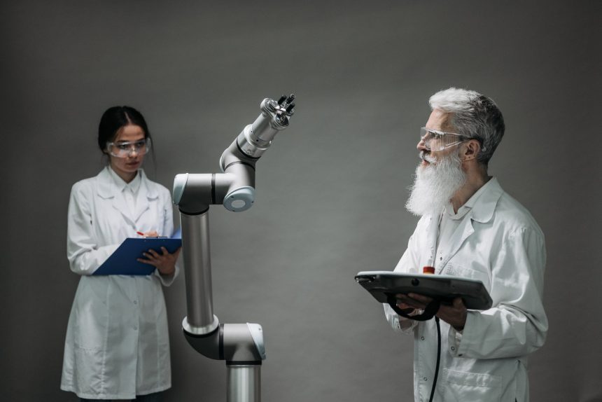 Dois pesquisadores de jaleco analisam um braço robótico em um laboratório.